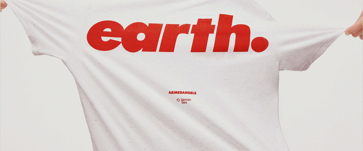 Nahaufnahme eines weißen T-Shirts mit großem roten Schriftzug ‚earth.‘ und kleinem Armedangels-Logo darunter; das Shirt wird seitlich gespannt gehalten.