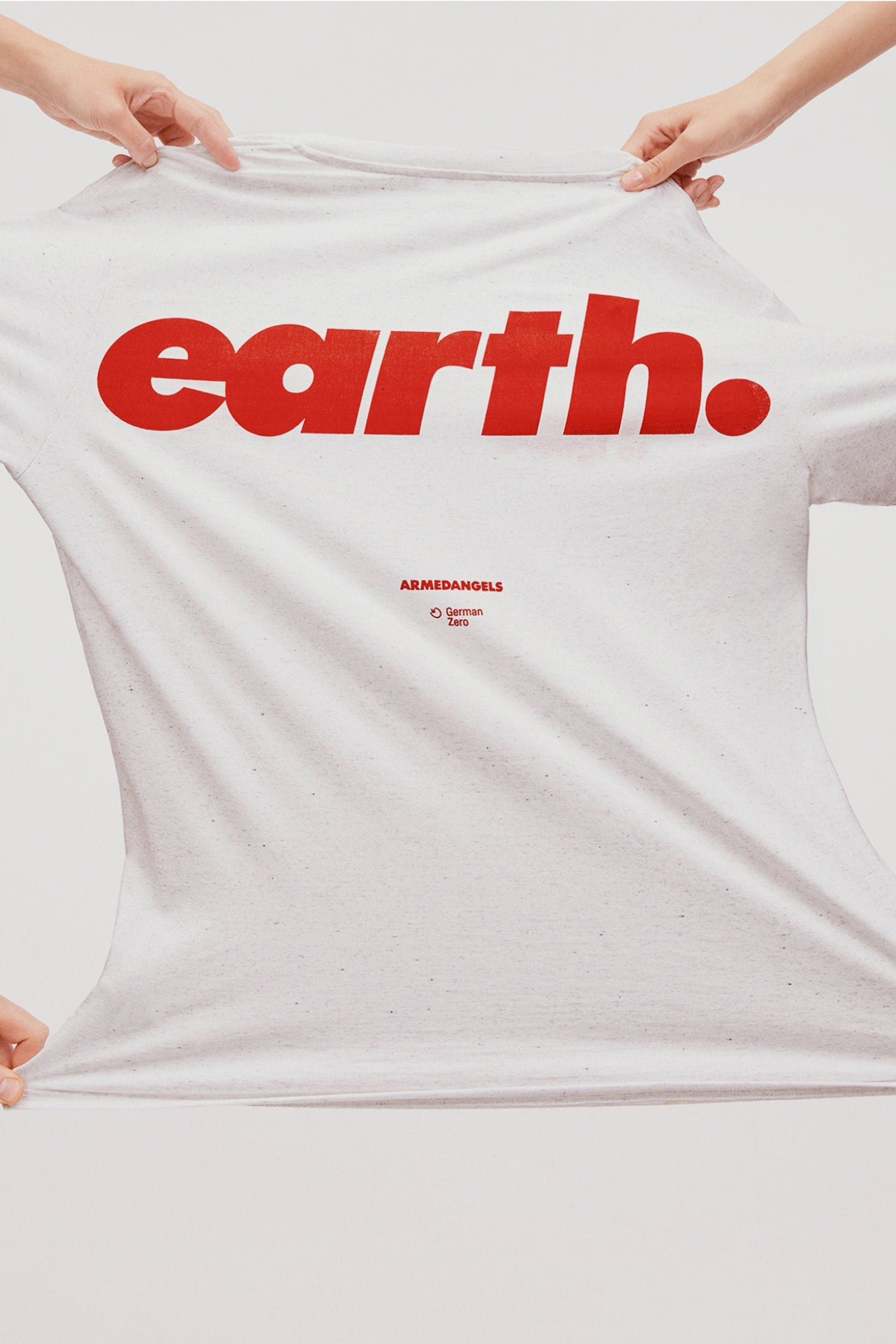 Nahaufnahme eines weißen T-Shirts mit großem roten Schriftzug ‚earth.‘ und kleinem Armedangels-Logo darunter; das Shirt wird seitlich gespannt gehalten.