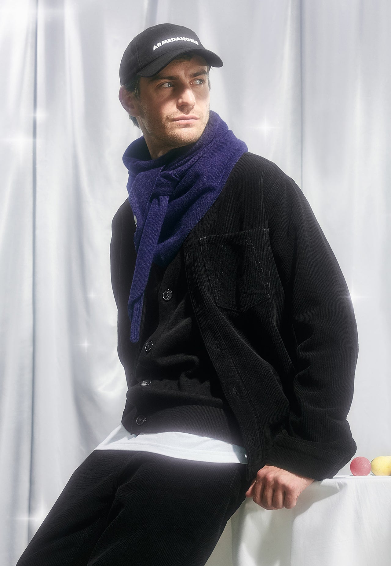 Eine Person trägt eine schwarze Cap, eine schwarze Cordjacke und einen violetten Schal und sitzt vor hellem, stofflichem Hintergrund.