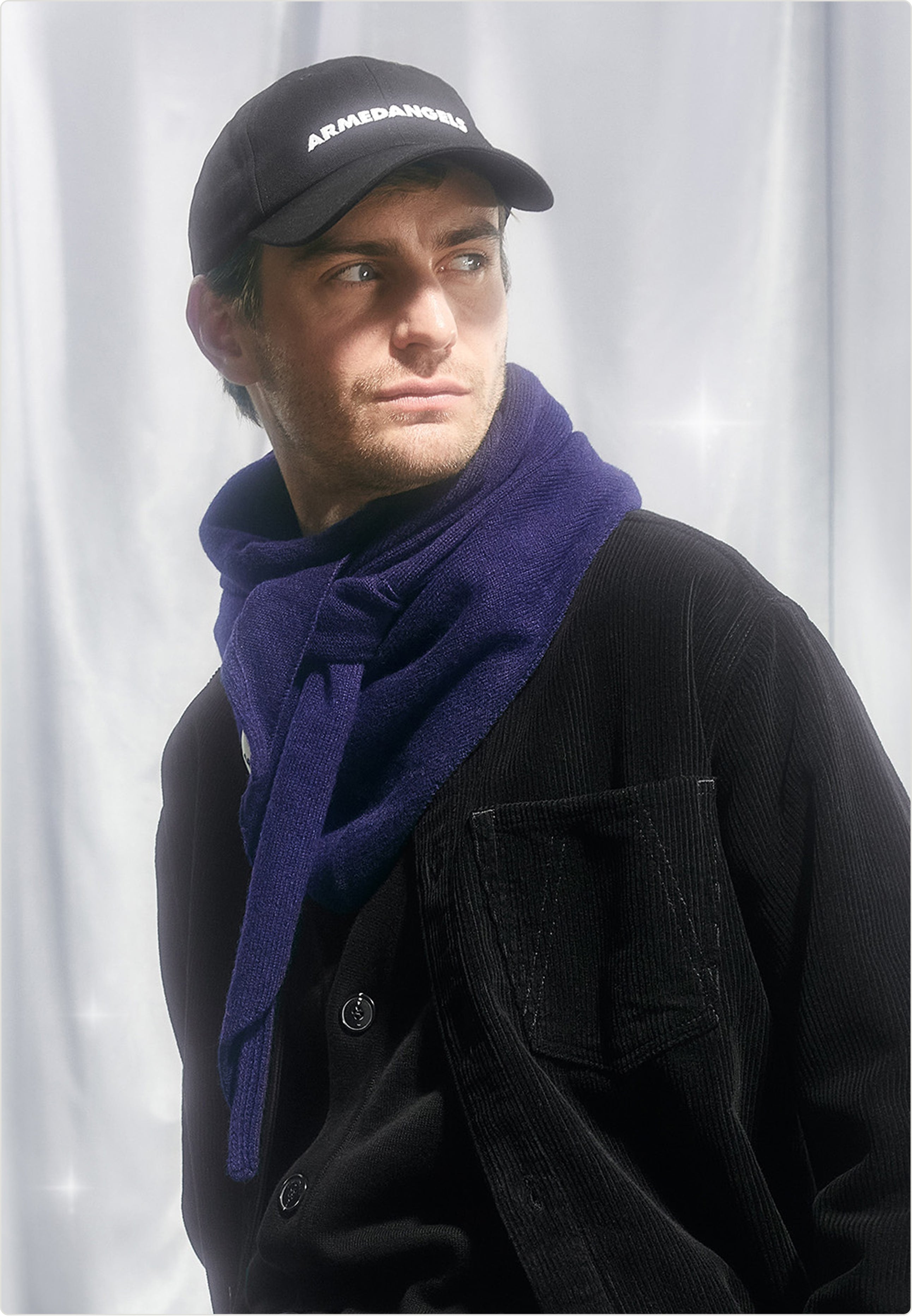 Eine Person trägt eine schwarze Cap, eine schwarze Cordjacke und einen violetten Schal und sitzt vor hellem, stofflichem Hintergrund.