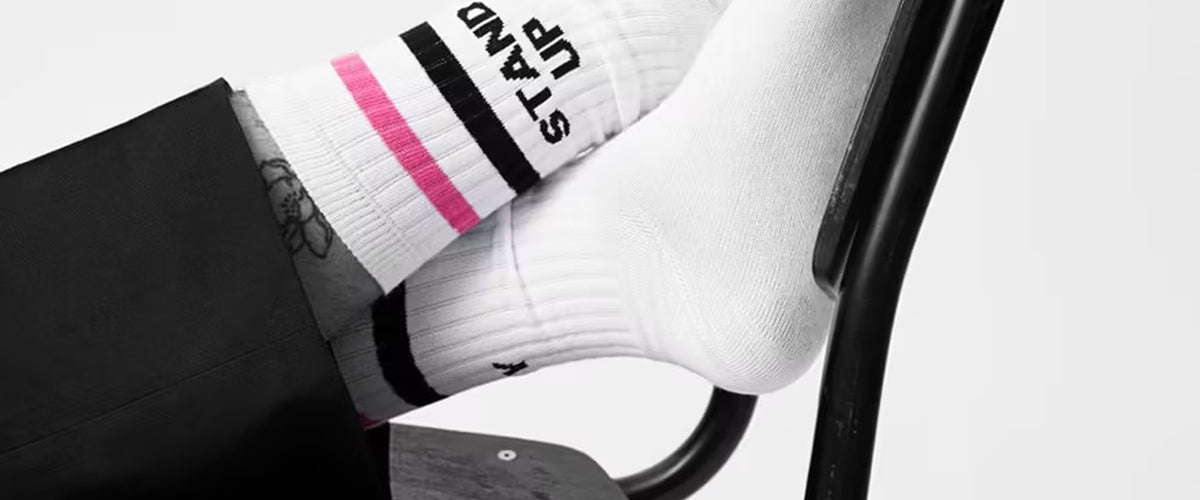Weiße Socken mit schwarzen und pinken Streifen und dem Schriftzug ‚STAND UP‘, getragen von einer Person, die die Füße auf eine schwarze Metallstuhlstrebe legt.