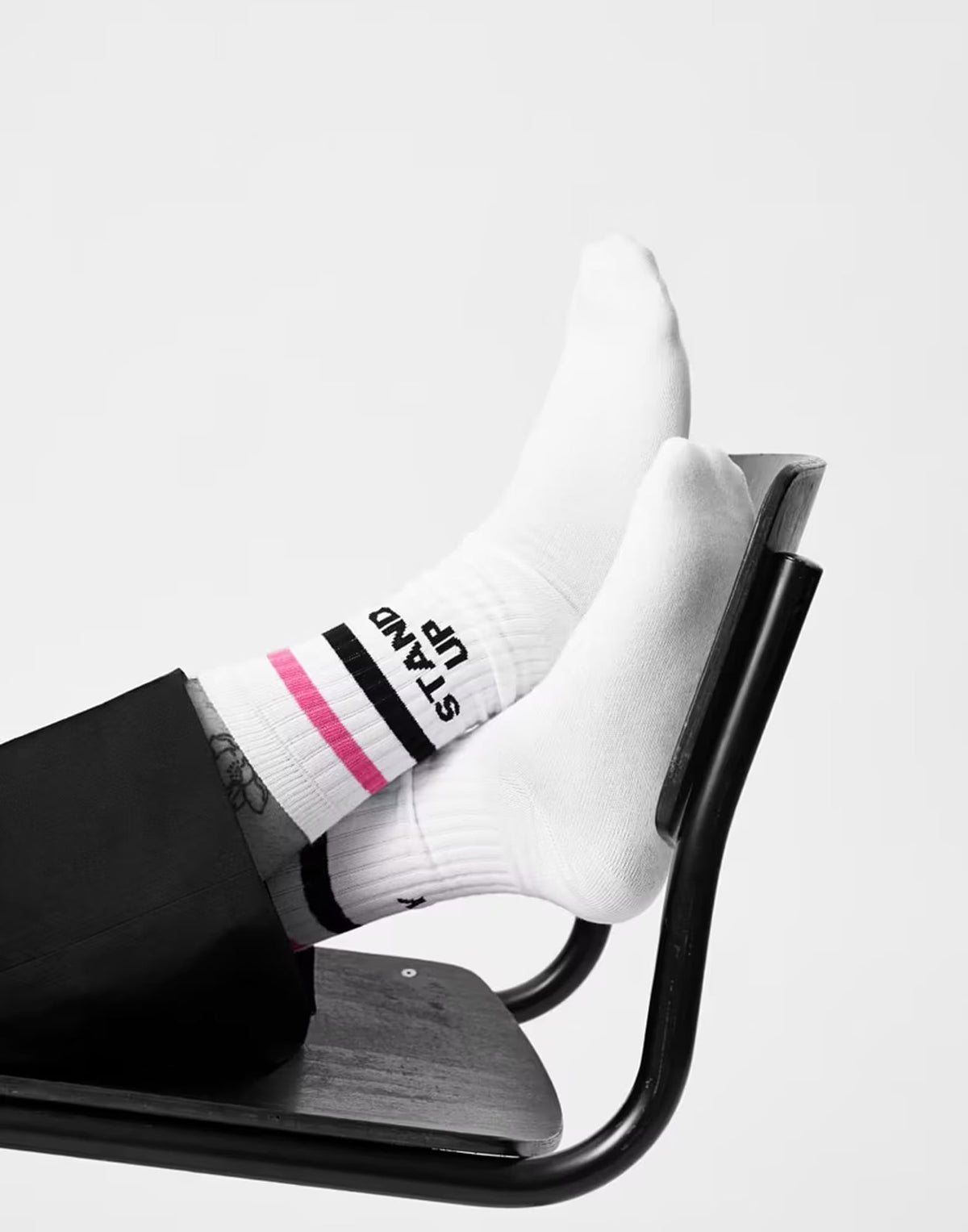 Weiße Socken mit schwarzen und pinken Streifen und dem Schriftzug ‚STAND UP‘, getragen von einer Person, die die Füße auf eine schwarze Metallstuhlstrebe legt.