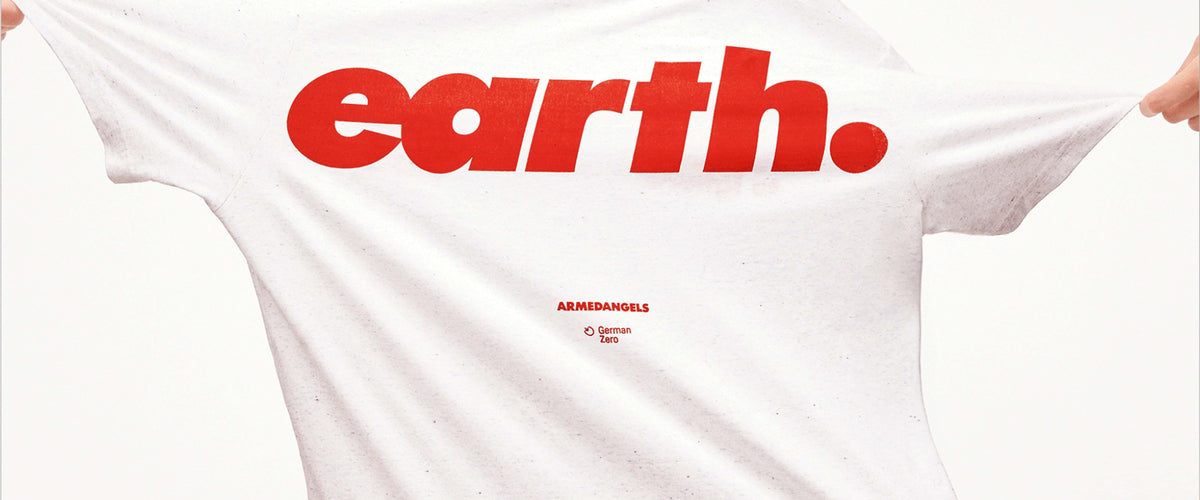 Nahaufnahme eines weißen T-Shirts mit großem roten Schriftzug ‚earth.‘ und kleinem Armedangels-Logo darunter; das Shirt wird seitlich gespannt gehalten.